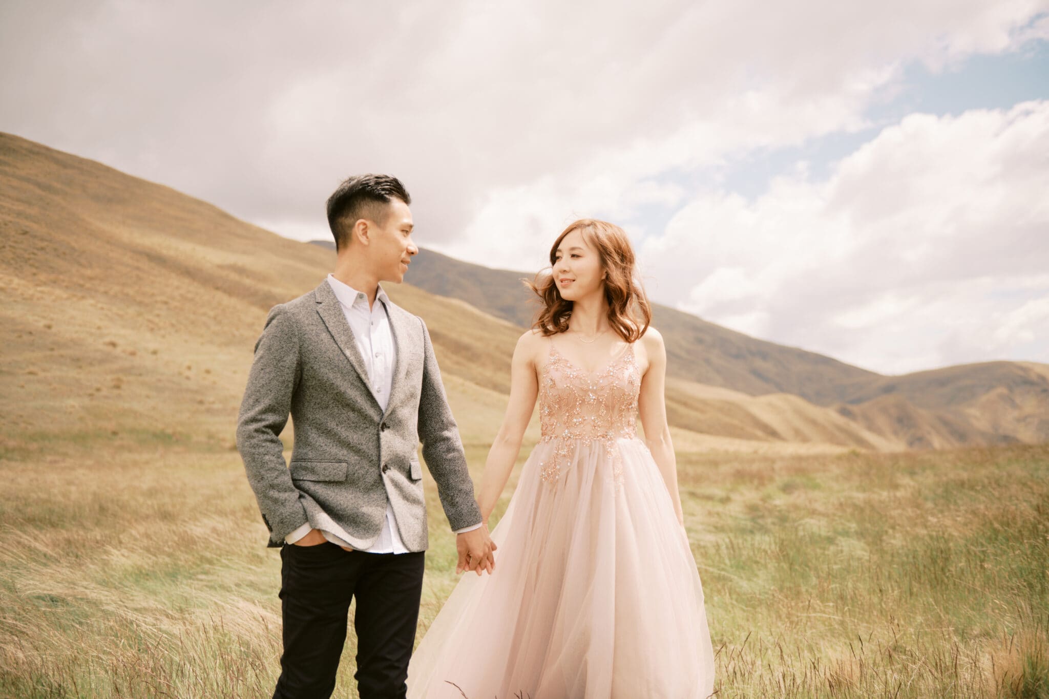 Queenstown Tekapo New Zealand Heli Wedding Elopement Pre-Wedding Shoot Photographer クイーンズタウン　テカポ　ニュージーランド　エロープメント 前撮り　フォトウェディング　結婚式 | A couple holding hands in a field with rolling hills in the background during their Tekapo pre-wedding shoot.